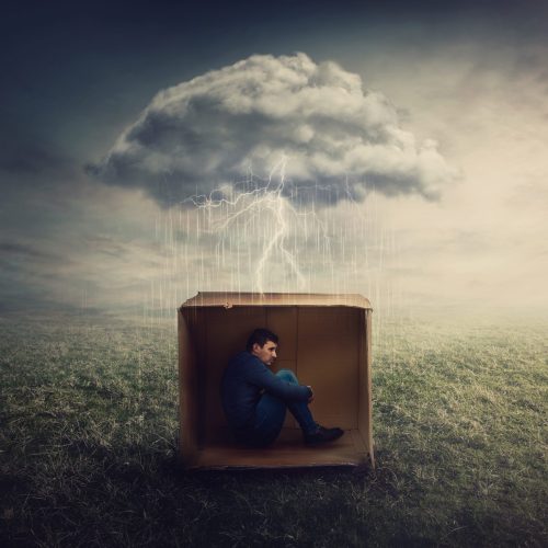 Surreal concept with a scared guy shelters inside a cardboard box. Introvert man caged by own fears as a thunderstorm cloud trapped him under the rain. Mysterious storm as emotional crisis symbol.