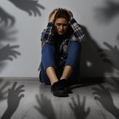 Paranoid delusion. Scared woman sitting near wall. Shadows of hands reaching for her symbolizing fear and anxiety